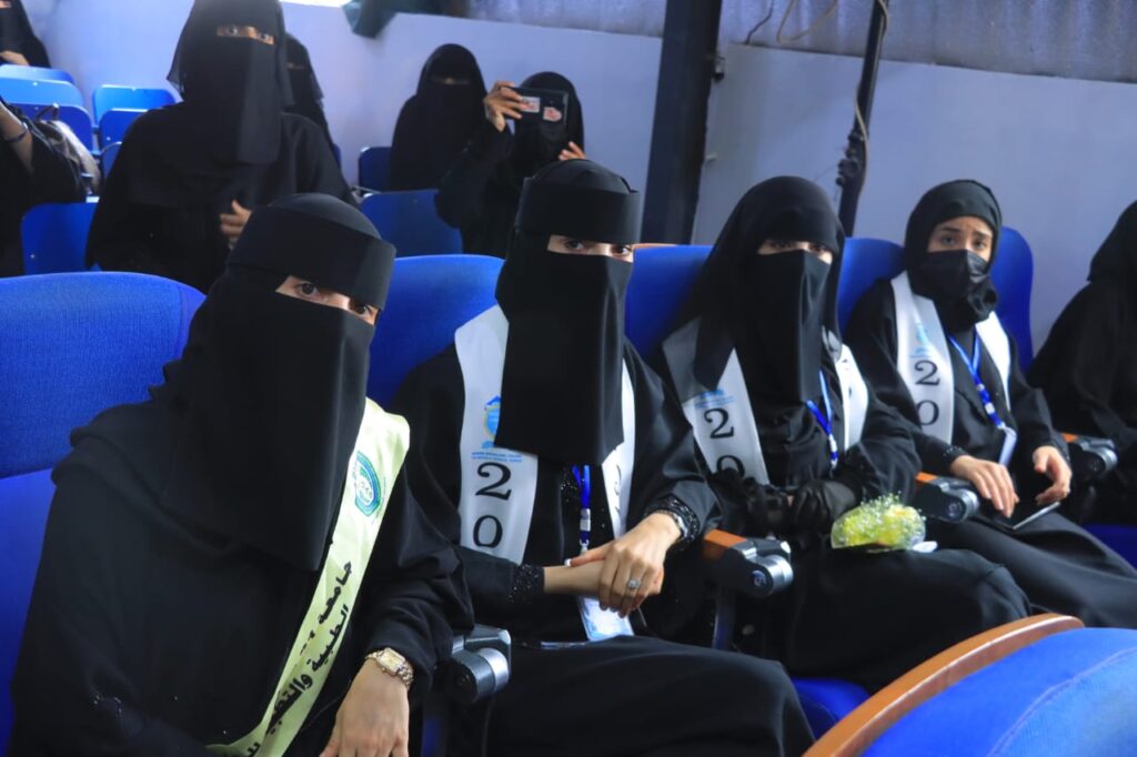 Women in black abayas and niqabs sit in blue theater seats at an event; sashes with the number 2020 are visible, one holds a bouquet and others have phones or cameras nearby.