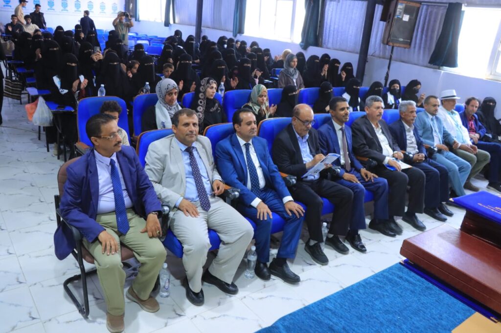 Front row of men in suits seated at a formal event; audience, mostly women in black abayas, sits in blue chairs behind them.