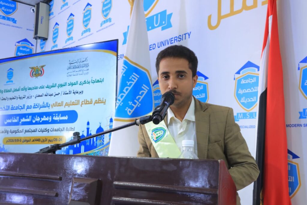 Young man in a beige suit speaks into a mic at a wooden podium during a university event, with Arabic banners and a flag in the background.