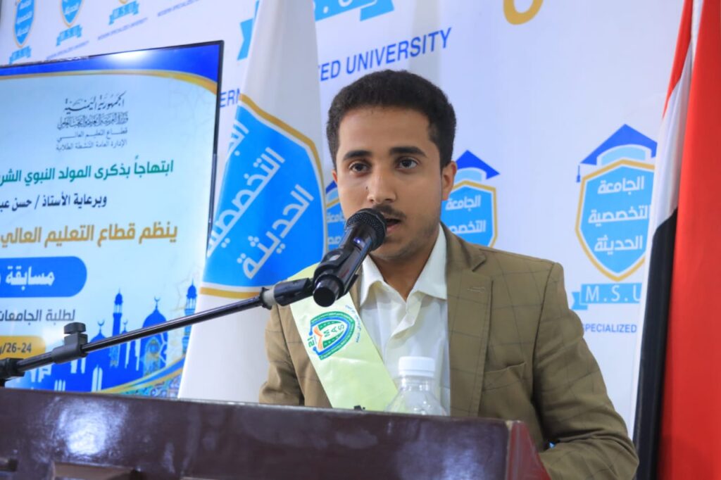 Young man in a tan blazer speaks into a microphone at a podium during a formal event, with a banner featuring Arabic text and university logos in the background (and a green sash across his chest).