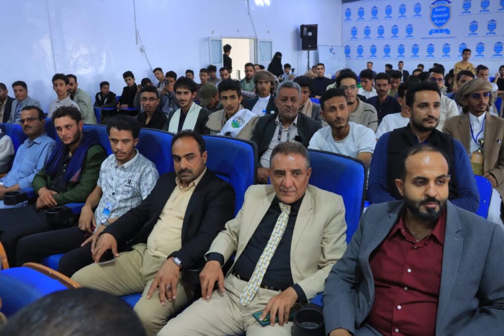 Audience of men seated in blue theater chairs at a conference, facing the stage.