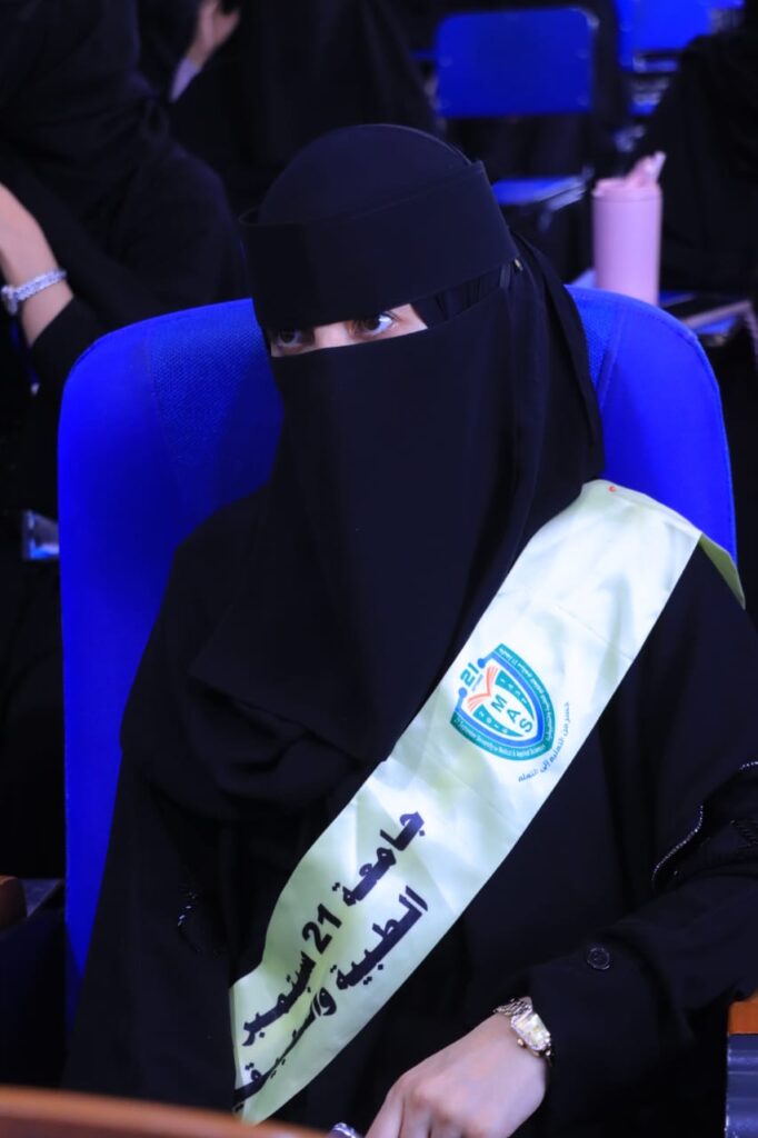 Woman in a black abaya and niqab seated in a blue chair, wearing a light green graduation sash and badge at a ceremony.