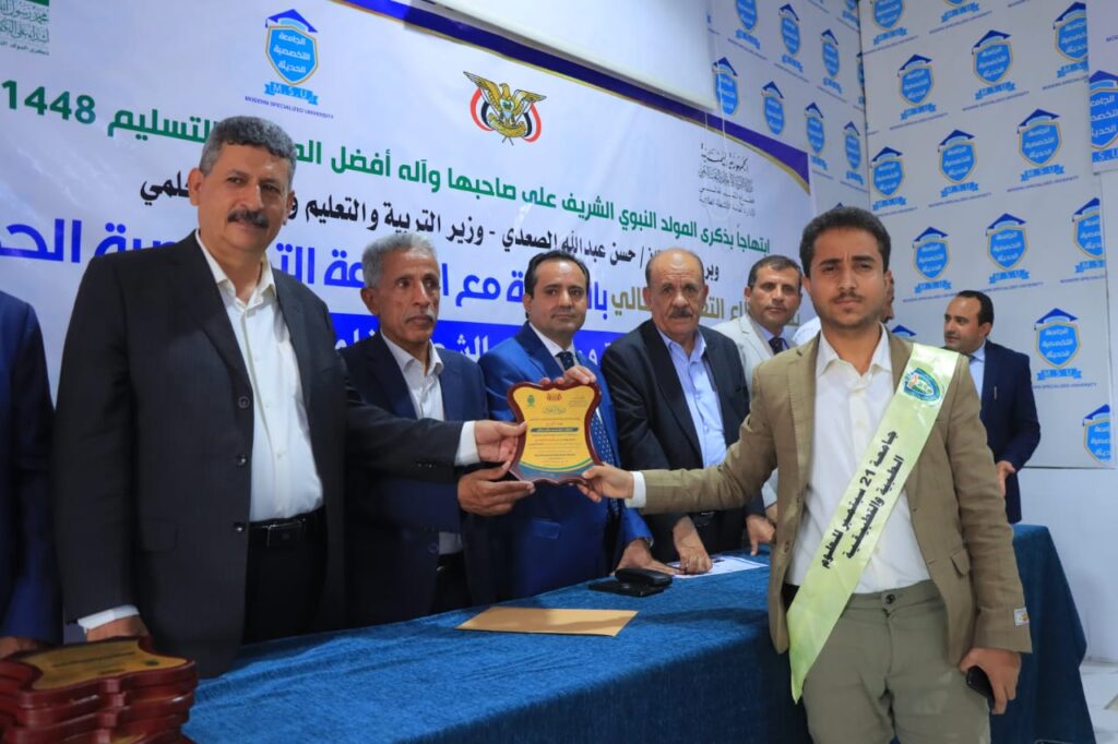 Award ceremony: a man in a beige suit with a sash receives a shield-shaped plaque from a colleague while other officials look on, with a blue table and Arabic banner in the background.