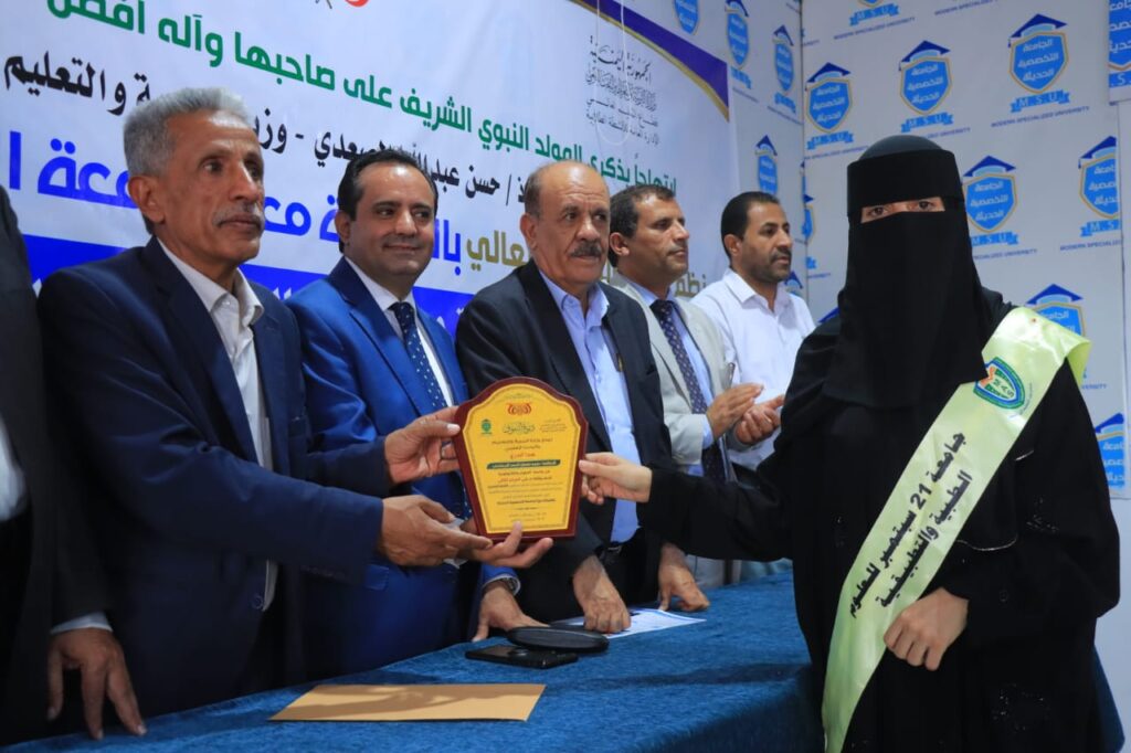 Officials in suits present a gold plaque to a woman in a black niqab and pale green sash at a ceremony by a blue-draped table.