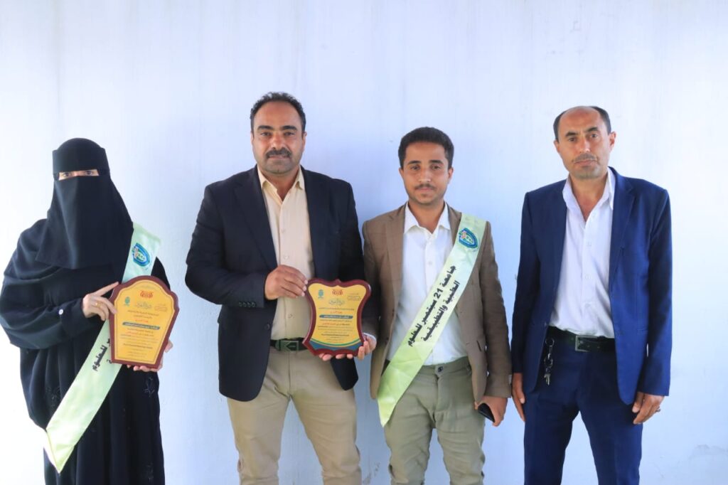 Four people stand against a pale blue wall; two men in suits hold plaques while a veiled woman and a young man wear light sashes, likely at an awards event.