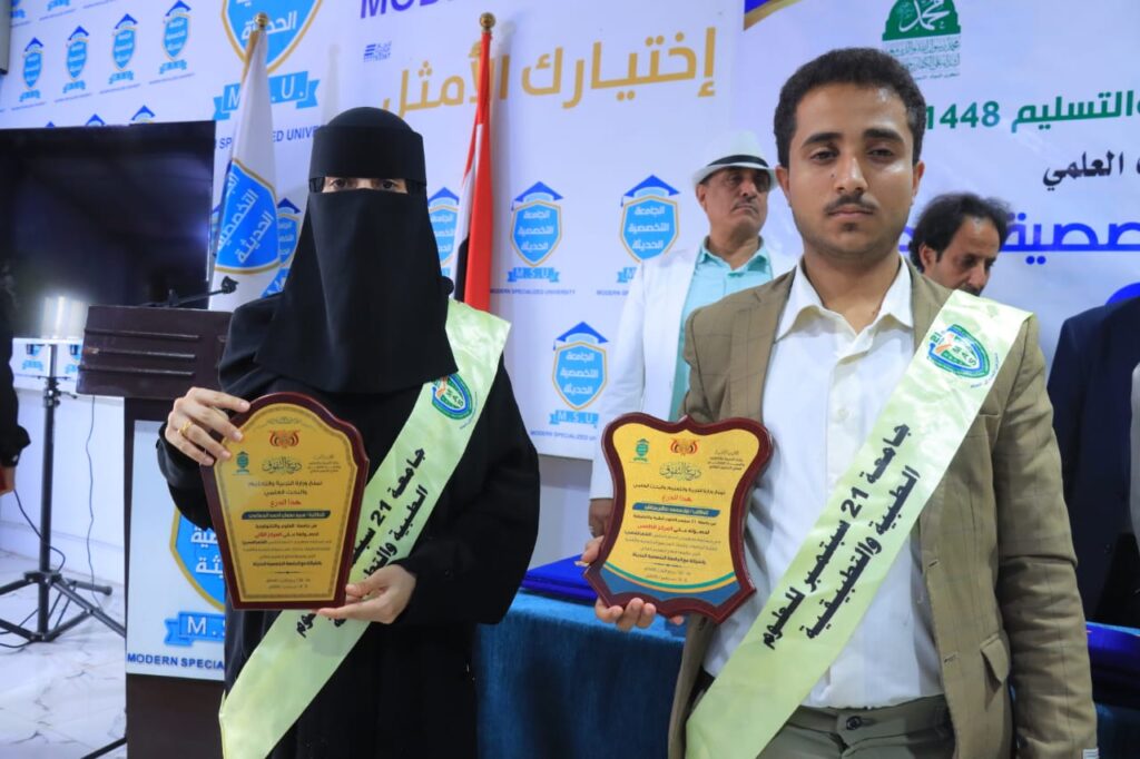 Woman in a niqab and a man in a suit hold shield-shaped plaques at an awards ceremony with a banner backdrop behind them.