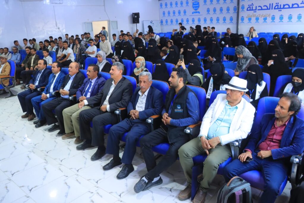 Front row of dignitaries in suits seated in a bright conference hall, with many attendees in blue seats and women in black abayas in the background.] ,