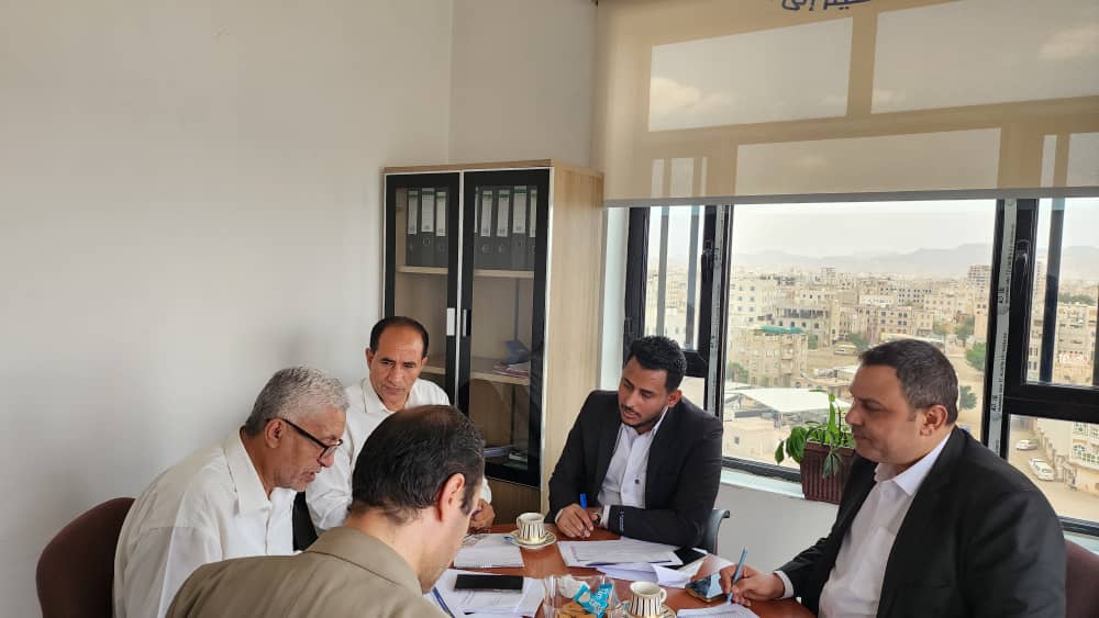 IMG-20260908-WA0119 - جامعة 21 سبتمبر Five men in business attire around a conference table in an office, reviewing documents with a city view through large windows.