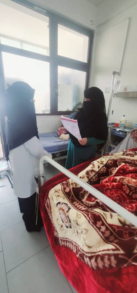 Nurse in a white coat and hijab consults with a patient in a hospital room; another patient lies in bed under a red patterned blanket near an IV pole.