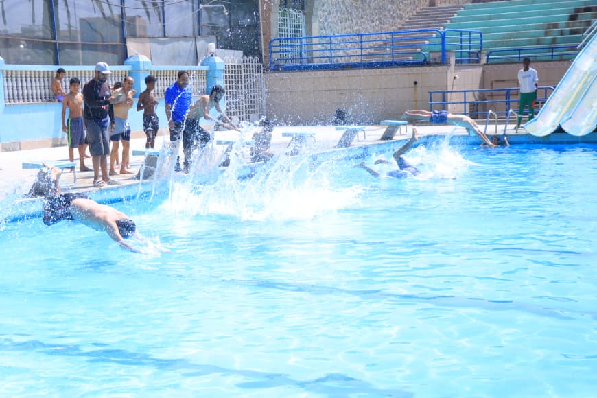 Children dive into a blue pool as supervisors watch nearby on a sunny day by the edge of the pool.