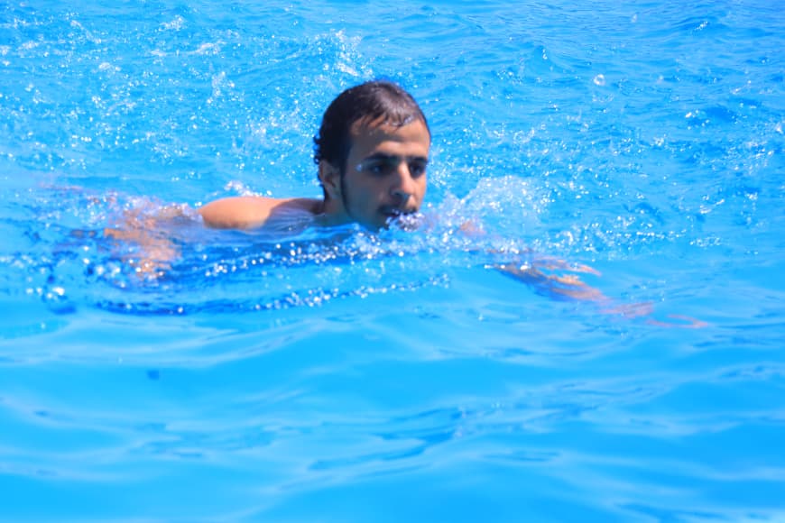 Man swimming in a bright blue pool, head above water, mid-stroke as he moves forward.