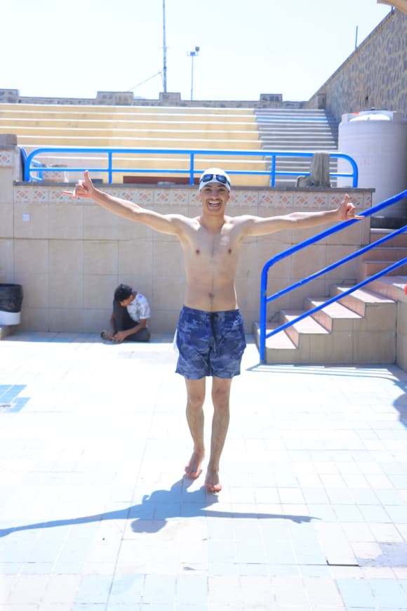 Young man in blue swim trunks and a cap poses with arms spread at an outdoor pool, sunny day and blue railings nearby.