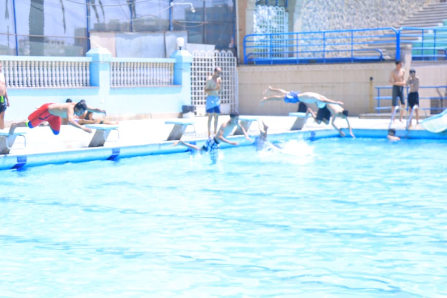 People dive jumps into a bright pool from the edge and diving boards; swimmers splash in the water.