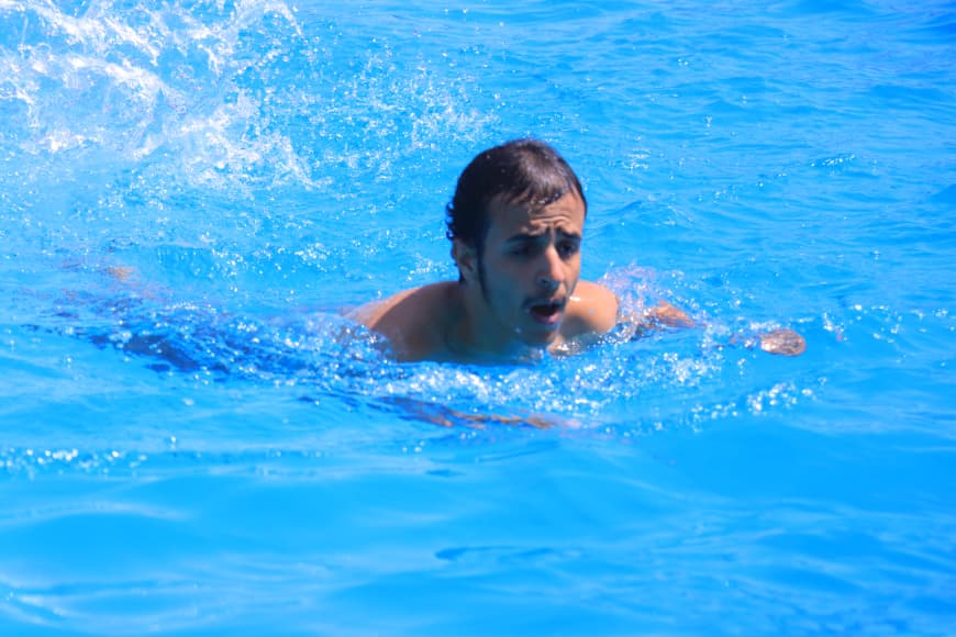 Man swimming in a bright blue pool, head and shoulders above water with splashing around him.