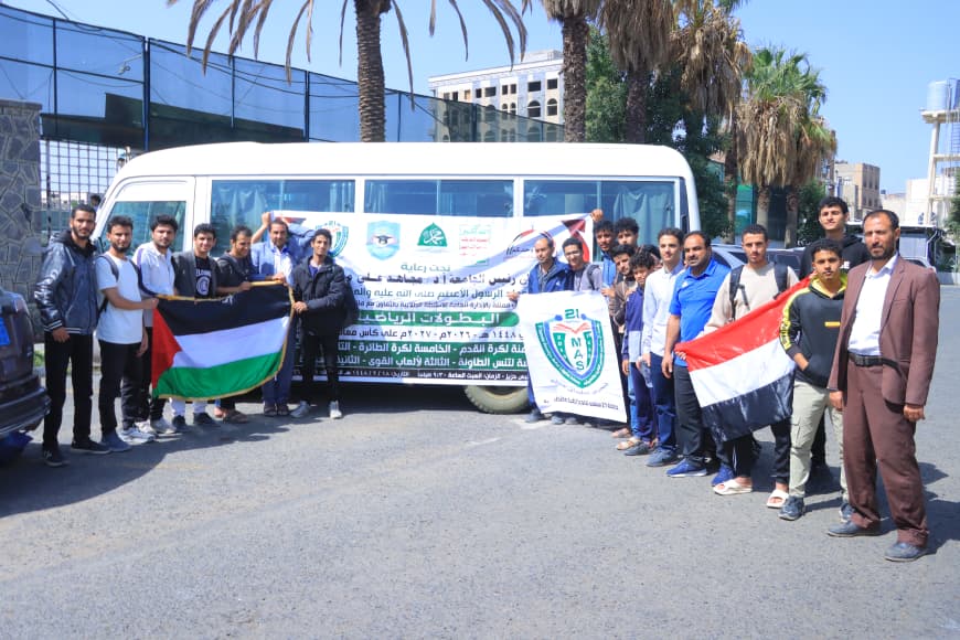 Group of men posing in front of a white bus, holding a large banner and flags (Palestinian flag) at a community event in a parking lot.