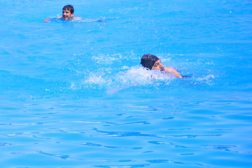 Two swimmers in a bright blue pool, one cutting through the water with splashes while another is visible in the background.