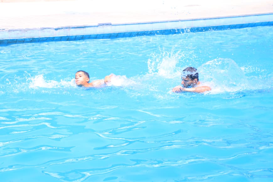 Two people swim in a bright blue pool near the edge, creating splashes as they paddle.