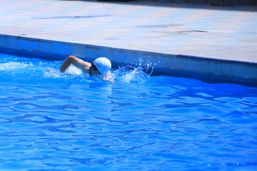 Swimmer in a bright blue outdoor pool wearing a white swim cap, arm extended in a freestyle stroke near the pool edge.