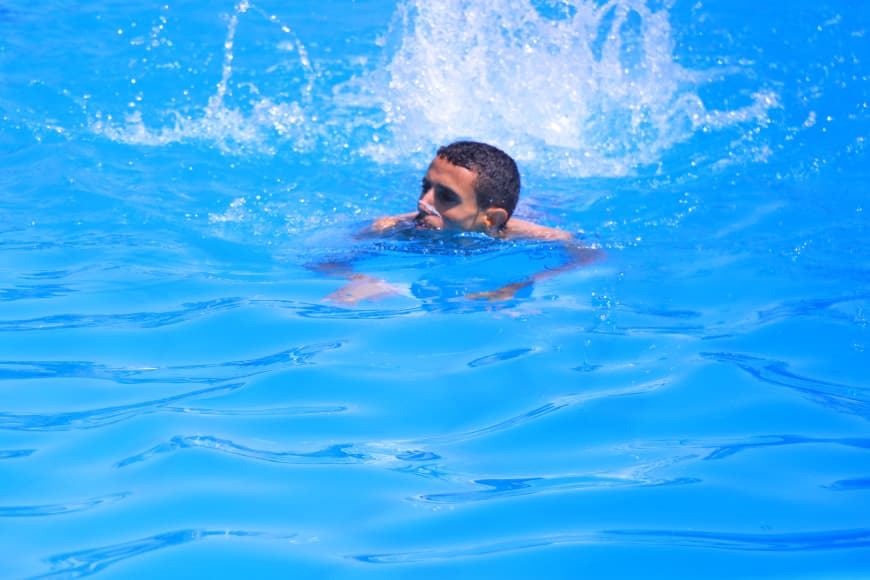 Man swimming in a bright blue pool, head above water with splashes behind him.