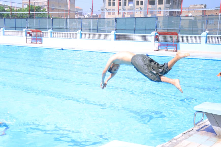 Man diving into an outdoor blue pool, mid-air, wearing dark swim trunks with pool deck and buildings in the background.
