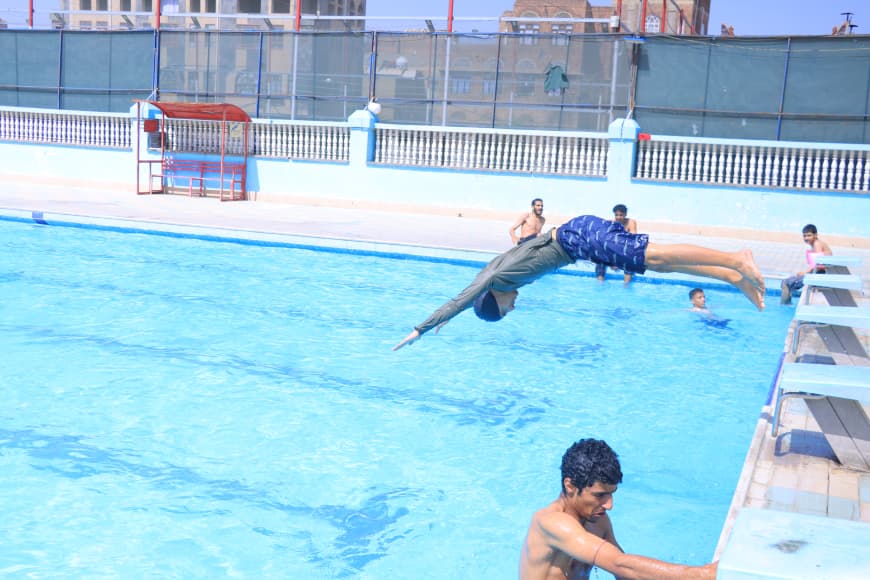 Two swimmers dive into a bright blue pool from the edge, while others watch from the poolside and a person sits at the far right.