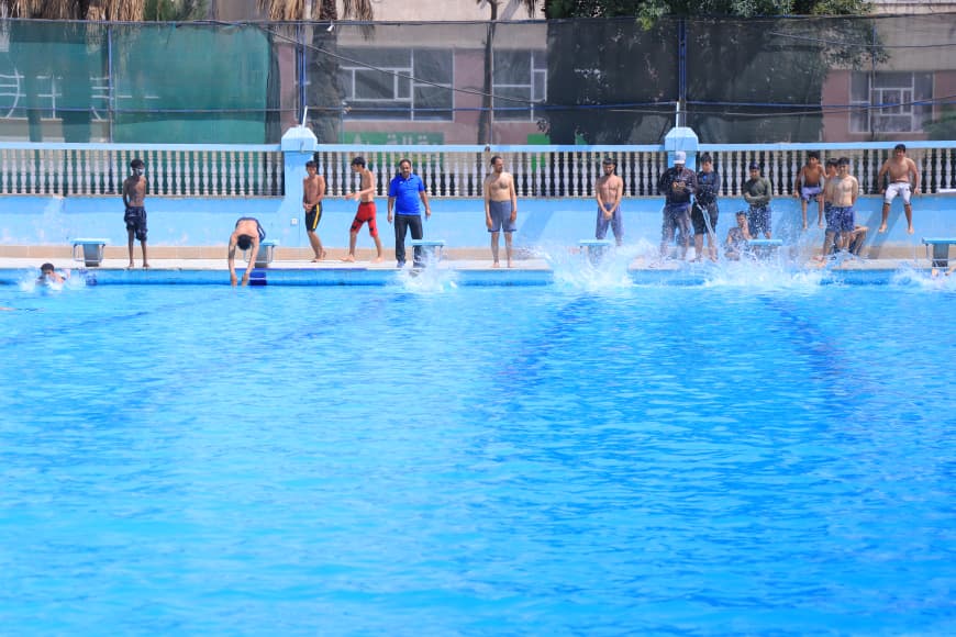 Group of people lined up along the edge of a bright blue swimming pool, with one person diving in and others watching as splashes occur in the water nearby.