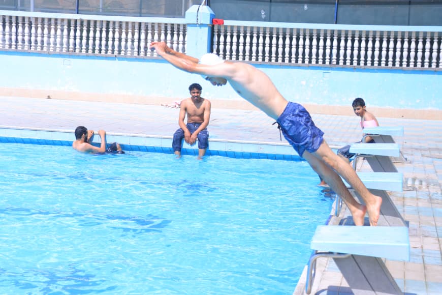 A man in blue swim trunks dives off a pool diving board into a bright blue outdoor pool, while others watch from the pool deck