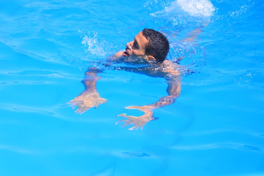 Man swimming in a bright blue pool with head above water and arms reaching forward