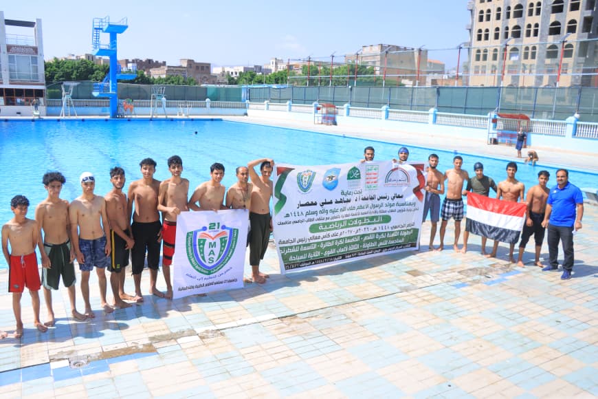 Group of young boys in swimsuits standing poolside, holding banners with logos; Egyptian flag on the right, city buildings in the background.