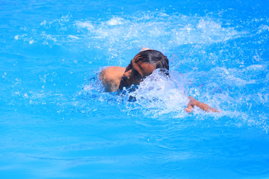 A swimmer in a bright blue pool, head and arm visible as water splashes around them.