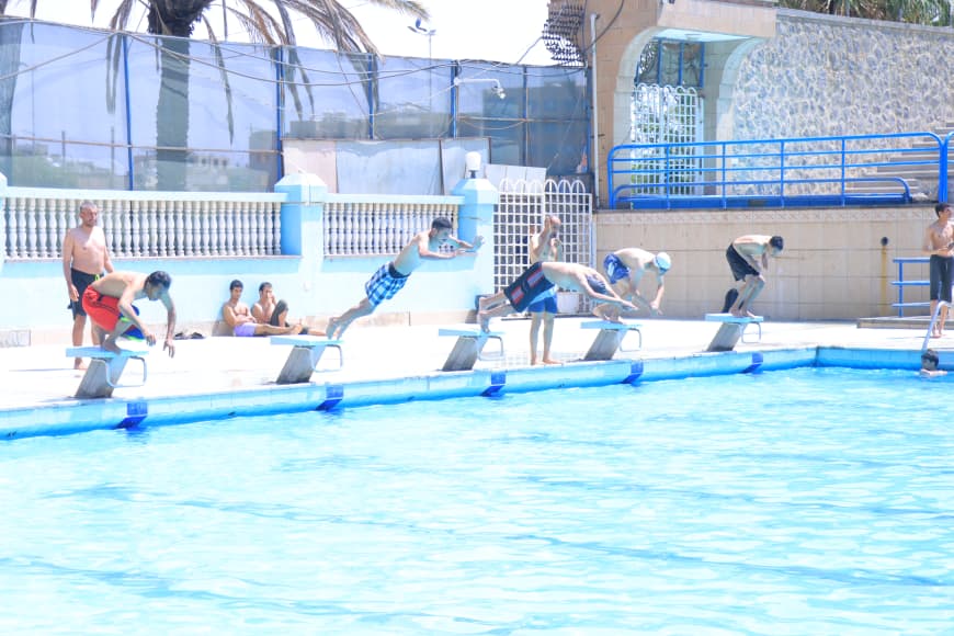 People dive from starting blocks into a bright outdoor pool, with a few observers nearby under sunny skies.
