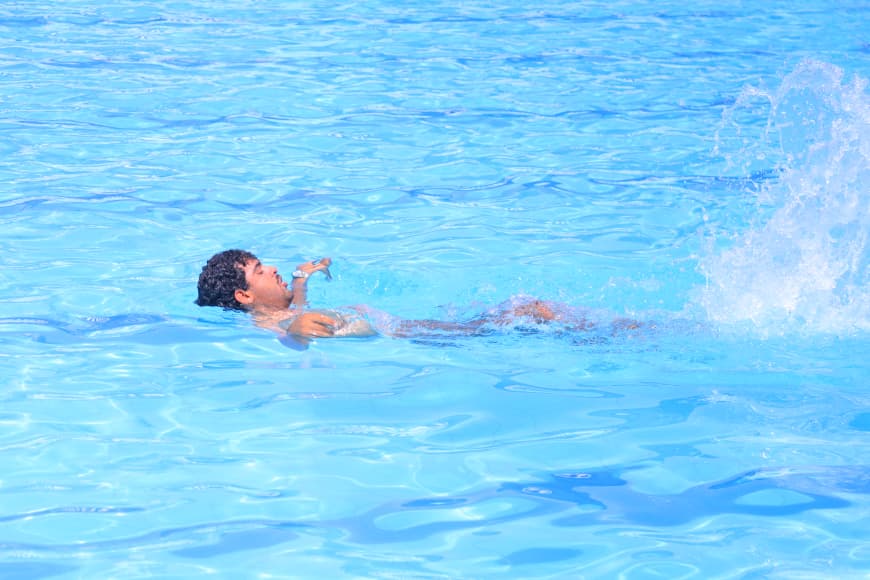 Man swimming on his back in a bright blue pool, creating a splash nearby at the surface.
