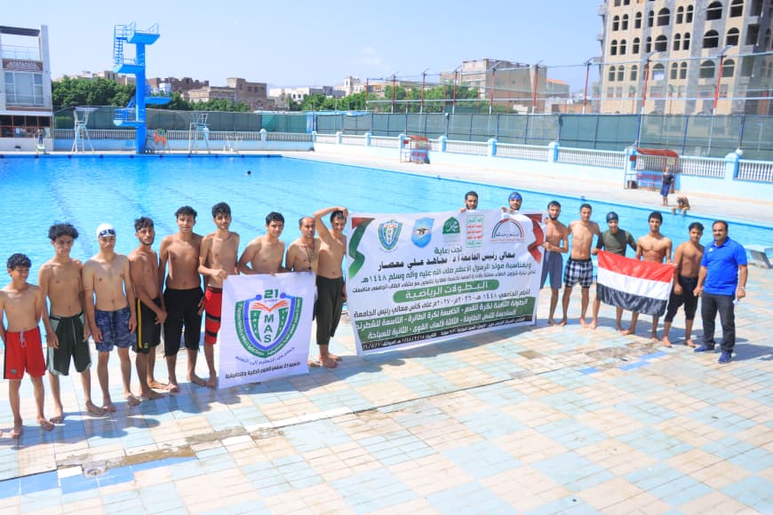 Group of shirtless boys standing at the edge of an outdoor swimming pool, holding banners and a red‑white‑black flag.