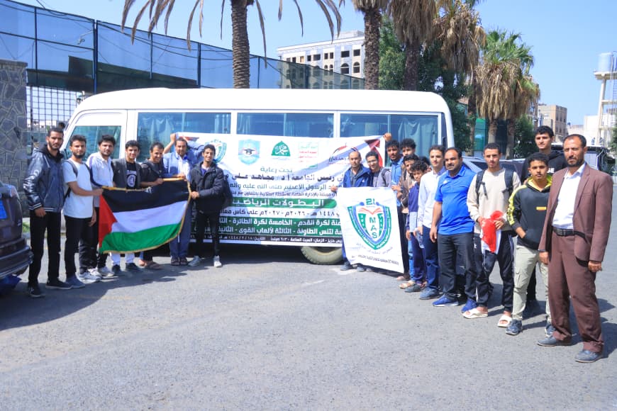 Group of men and a few adults stand beside a white bus, holding a Palestinian flag and a large banner with Arabic text for a community event.