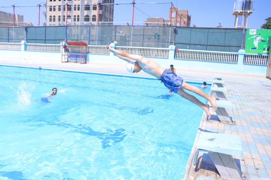 A swimmer wearing a white cap dives headfirst from the pool edge into the blue water as others swim nearby.