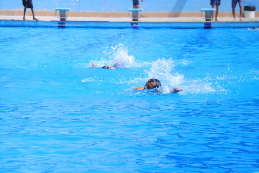 Two swimmers in a bright blue outdoor pool creating splashes near the surface; pool edge and bystanders visible in the background.