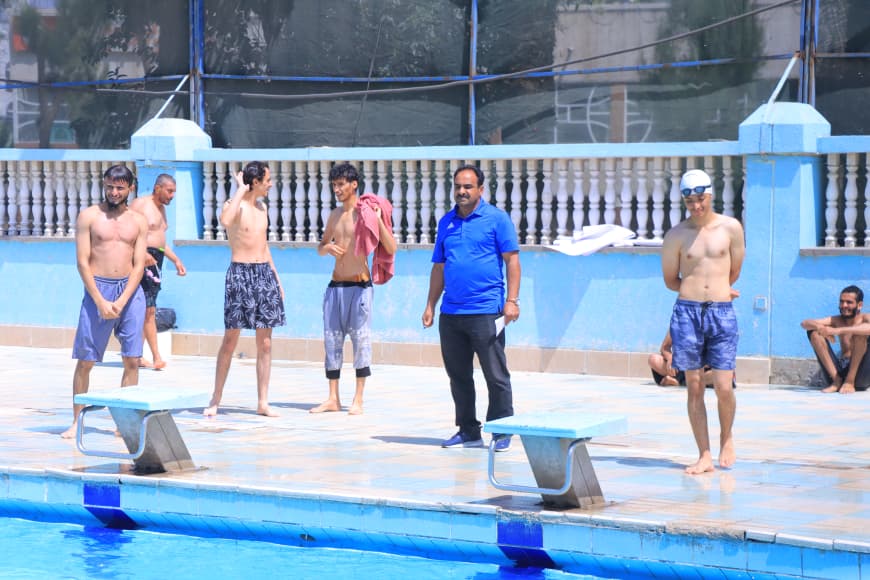 Poolside scene with young swimmers and a man in a blue polo standing by diving blocks by the pool edge.