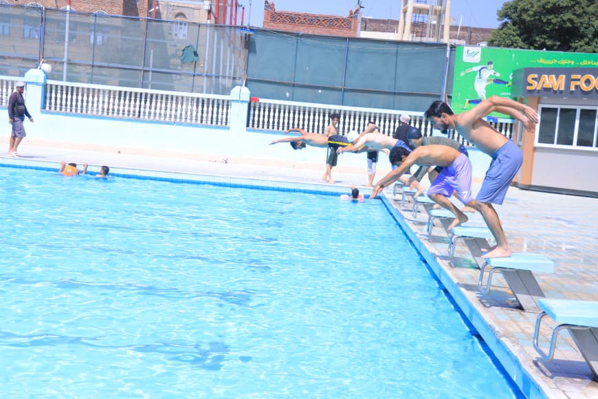 People dive from starting blocks into a bright outdoor pool as others watch along the pool deck.
