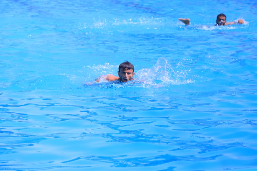 Two people swimming in a bright blue pool, one in the foreground and one in the distance.