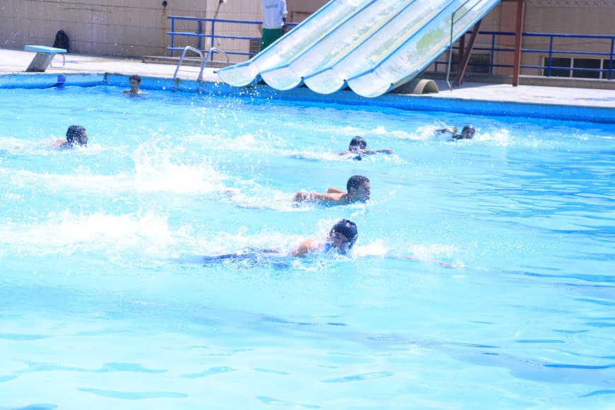 Several children swim in a bright blue pool with a large water slide in the background at a recreational facility.