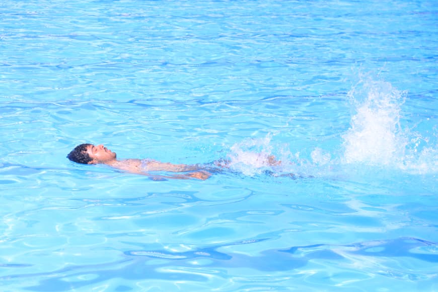 Person floating and kicking on their back in a bright blue pool, creating a splash behind them