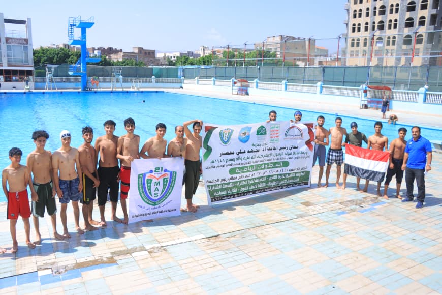 Group of young male swimmers standing barefoot along a pool edge, holding banners from a sports event or team gathering.