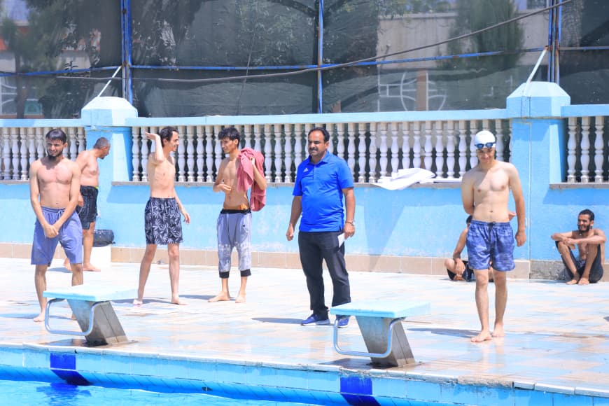 Poolside group of shirtless men listening to a man in a blue polo who appears to be coaching.