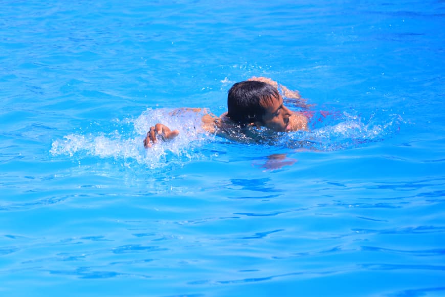 A person swimming in a bright blue pool, head turned to the side and arms splashing through the water."