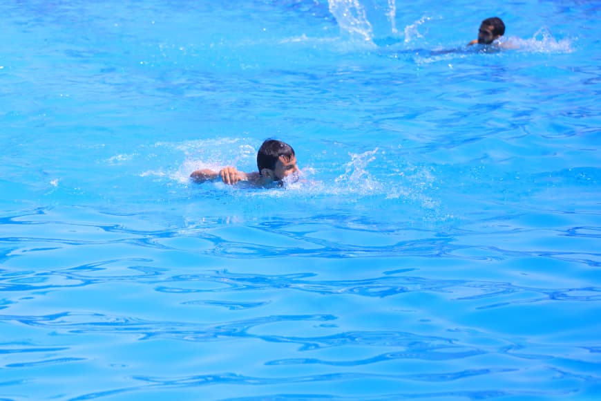 Two people swim in a bright blue pool, one in the foreground paddling with an arm forward while another swimmer splashes in the background
