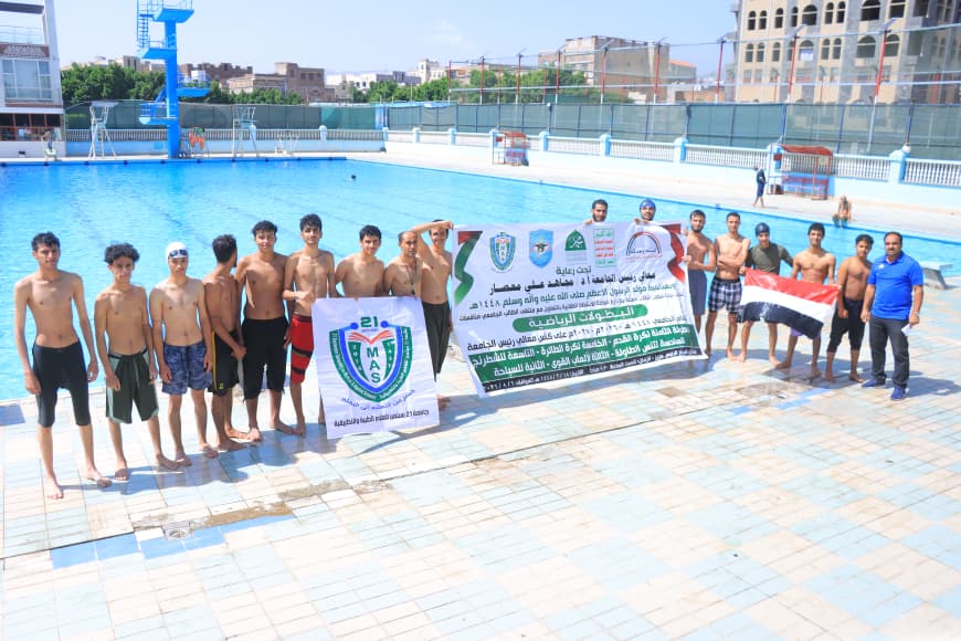 Group of shirtless young men standing at the edge of an outdoor swimming pool, holding banners and a flag.