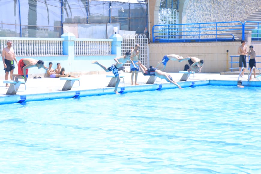 Outdoor pool scene with several youths leaping from diving boards into bright blue water while onlookers sit along the pool edge.