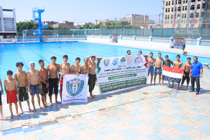 Group of young swimmers standing in a row by a large outdoor pool, holding banners and an Egyptian flag.