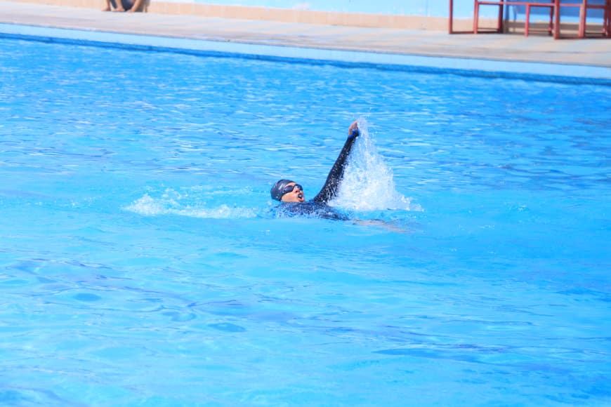 Swimmer in a bright blue outdoor pool raises one arm above the water during a stroke, with the pool edge visible in the background.