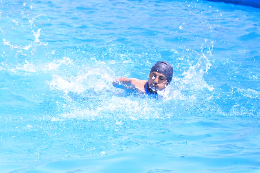Swimmer wearing a dark swim cap glides through a bright blue pool with splashing water around.
